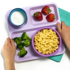Hands holding a Purple school tray with instant pot Mac and cheese, broccoli and strawberries for a toddler or kid.