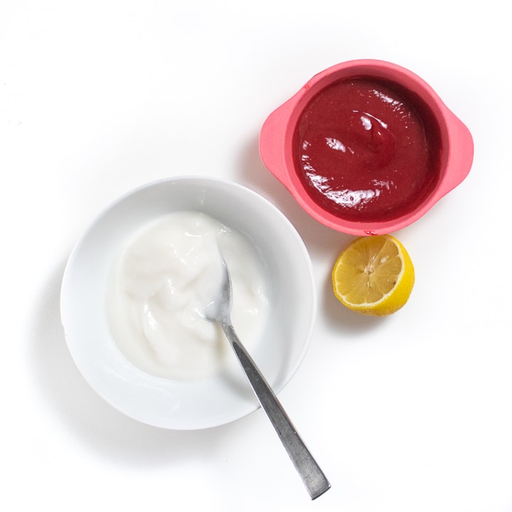 White bowl with yogurt, lemon and a pink bowl of raspberry puree on a white background.