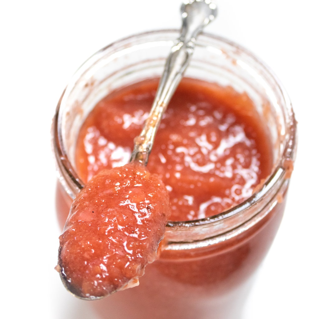 A jar full of strawberry applesauce with a silver spoon on top filled with strawberry applesauce.