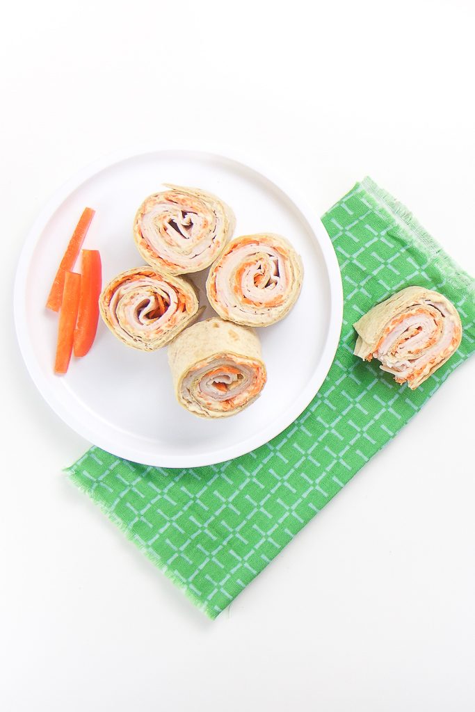 a small white plate sitting on a green napkin on top of a white surface. Place has 4 turkey carrot rollups with 3 slices of red peppers on the plate. On the napkin is one half eaten rollup.