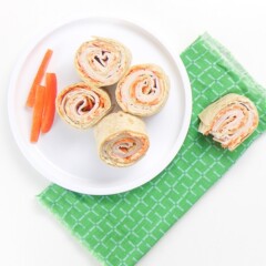 a small white plate sitting on a green napkin on top of a white surface. Place has 4 turkey and carrot rollups wit 3 slices of red peppers on the plate. On the napkin is one half eaten rollup.